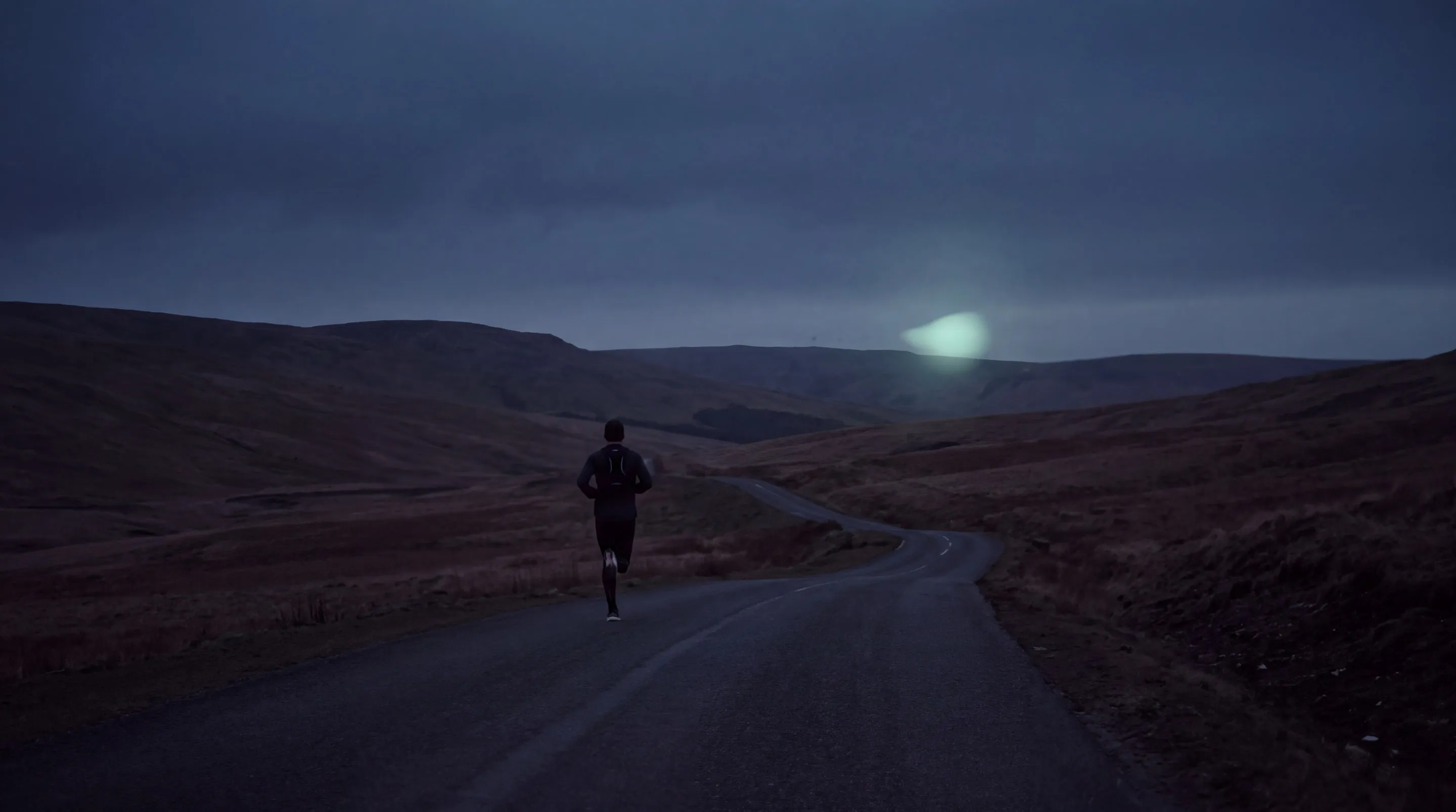 Lone runner on a winding moorland road at dusk, a soft mint light glowing above the hills